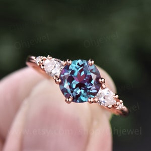 Pode incluir: Um anel de ouro rosa com uma grande gema redonda, azul e roxa iridescente, cercada por gemas menores e transparentes.