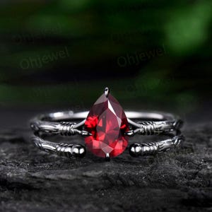 May include: A pear-shaped red gemstone ring with a black metal band. The band is twisted and has a delicate design. The ring is set on a dark background.