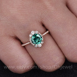 May include: A pear-shaped emerald engagement ring with a halo of small round diamonds set in rose gold. The ring is on a finger with a white background.