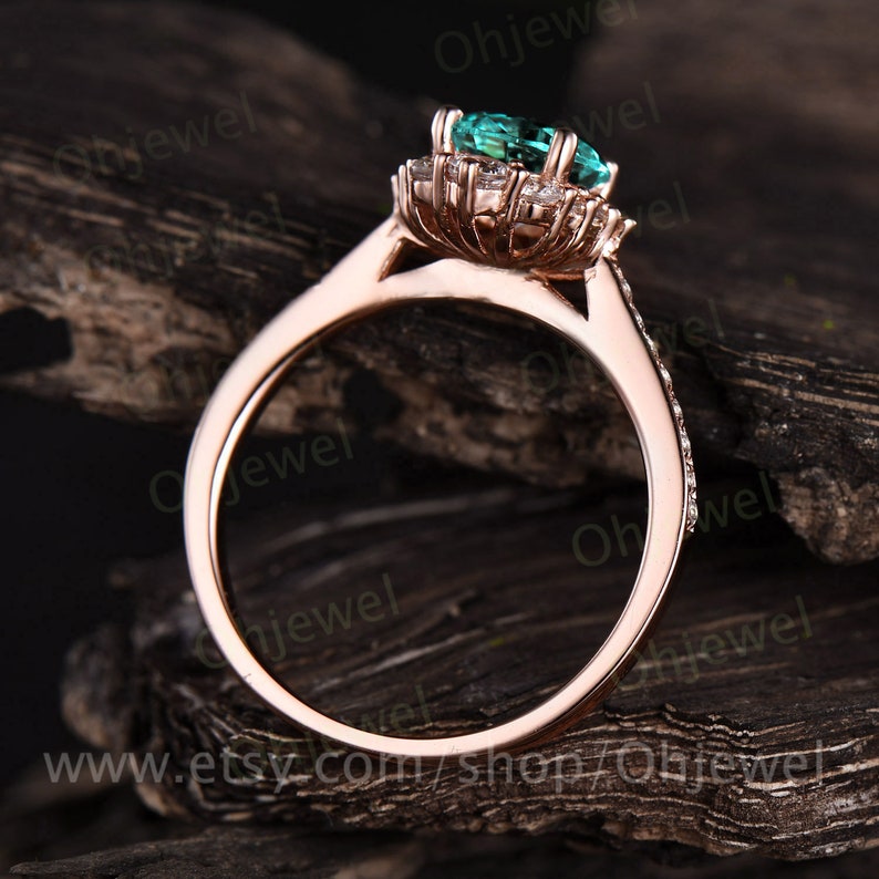 May include: A rose gold engagement ring with a green oval gemstone surrounded by a halo of smaller diamonds. The ring is set on a wooden surface.
