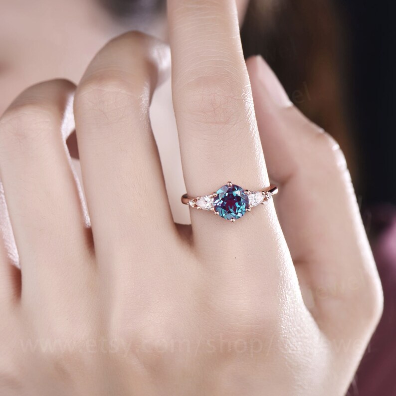 Pode incluir: Um anel de ouro rosa com uma gema redonda azul-esverdeada cercada por tr&ecirc;s gemas brancas menores em forma de p&ecirc;ra.