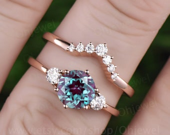 May include: A rose gold ring set with a large, green and blue alexandrite gemstone surrounded by smaller diamonds. The set includes a matching rose gold band with a curved row of diamonds.