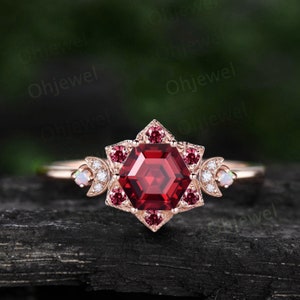 May include: A rose gold engagement ring with a hexagonal red gemstone surrounded by smaller red gemstones and diamonds. The ring also features two crescent moon accents with opal and diamond stones.