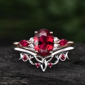 May include: A ruby engagement ring set with a large oval-shaped ruby surrounded by smaller diamonds. The ring is set in a delicate, ornate band with a Celtic knot design. The ring is paired with a matching wedding band with a smaller ruby and diamonds.