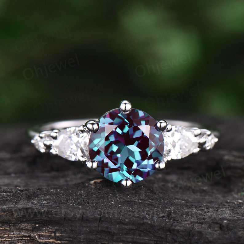 Pode incluir: Um anel de prata com uma grande gema redonda e iridescente azul e roxa, cercada por tr&ecirc;s gemas menores em forma de p&ecirc;ra, transparentes.