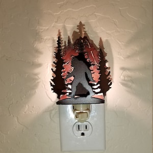 May include: A metal night light featuring a silhouette of Bigfoot walking through a forest. The night light is plugged into a standard electrical outlet.