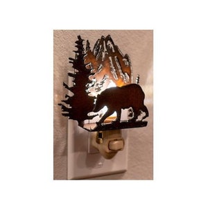 May include: A metal night light shaped like a bear walking through a forest with mountains in the background. The night light is plugged into an electrical outlet and is lit up.