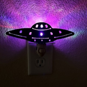 May include: A black UFO-shaped night light plugged into a standard electrical outlet. The light is on and emitting a purple glow.