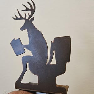 May include: A dark brown metal night light featuring a silhouette of a deer with antlers sitting on a toilet while reading a book. The decorative cutout design is mounted on a standard white plastic electrical plug-in base with a small toggle switch.