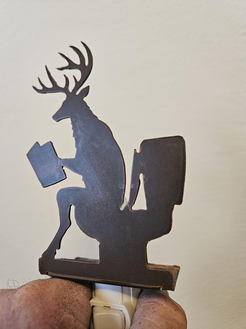 May include: A dark brown metal night light featuring a silhouette of a deer with antlers sitting on a toilet while reading a book. The decorative cutout design is mounted on a standard white plastic electrical plug-in base with a small toggle switch.