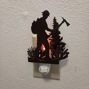 May include: A dark brown metal night light featuring a silhouette of a firefighter wearing a helmet and carrying an axe, standing in front of a pine tree. The light glows from behind the metal cutout, illuminating the scene from the wall outlet.