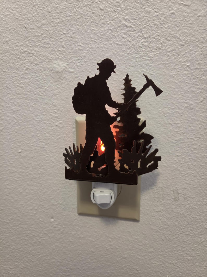 May include: A dark brown metal night light featuring a silhouette of a firefighter wearing a helmet and carrying an axe, standing in front of a pine tree. The light glows from behind the metal cutout, illuminating the scene from the wall outlet.