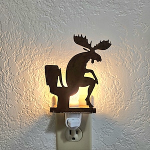 May include: A dark brown metal night light featuring a silhouette of a moose sitting on a toilet. The light is plugged into a wall outlet, emitting a warm glow from behind the metal cutout, illuminating the wall around the fixture.