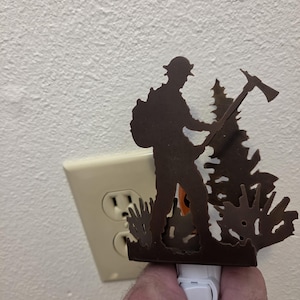 May include: A dark brown metal night light featuring a silhouette of a firefighter wearing a helmet and carrying an axe, standing next to a pine tree. The decorative cover is attached to a white plastic plug-in base with a small toggle switch.