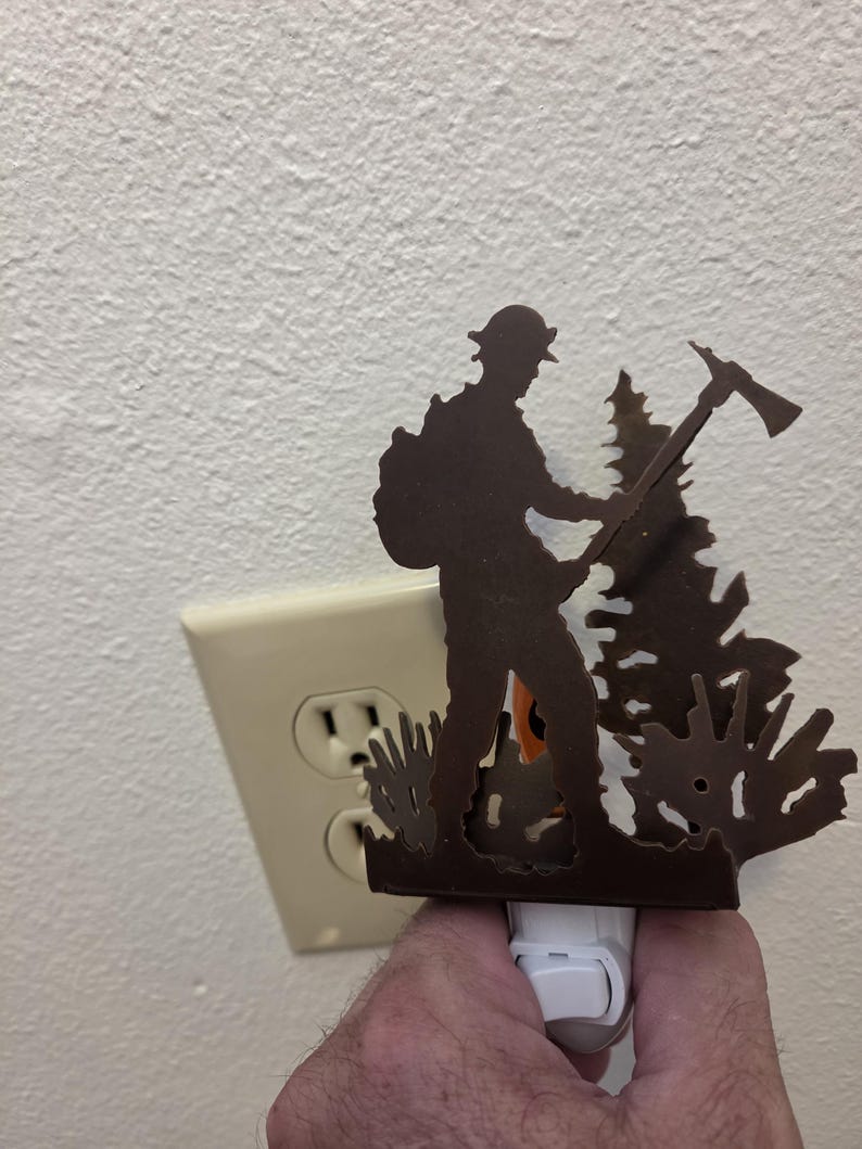 May include: A dark brown metal night light featuring a silhouette of a firefighter wearing a helmet and carrying an axe, standing next to a pine tree. The decorative cover is attached to a white plastic plug-in base with a small toggle switch.