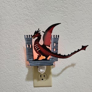 May include: A red dragon night light with a gray castle design. The night light is plugged into an electrical outlet.