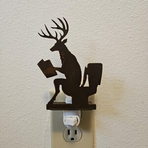 May include: A dark brown metal night light featuring a silhouette of a deer with antlers sitting on a toilet while reading a book. The decorative piece is mounted onto a standard white electrical wall plug-in base with a small toggle switch.