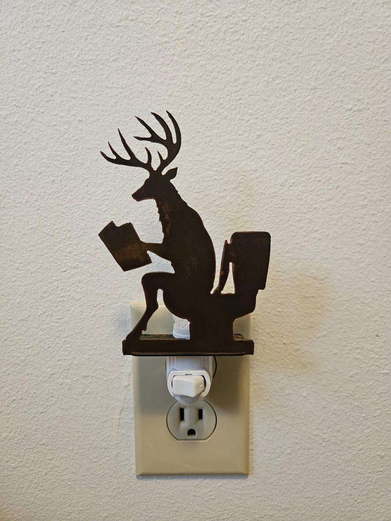 May include: A dark brown metal night light featuring a silhouette of a deer with antlers sitting on a toilet while reading a book. The decorative piece is mounted onto a standard white electrical wall plug-in base with a small toggle switch.