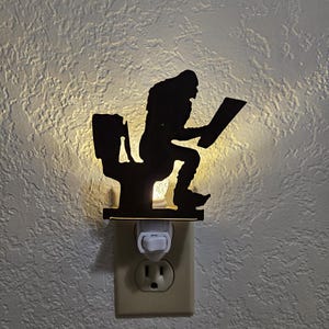 May include: A black silhouette night light featuring a figure seated on a toilet, reading. The night light plugs into a standard wall outlet. The light shines from behind the figure, creating a shadow effect. The wall is textured.