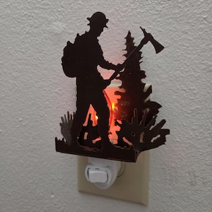 May include: A dark brown metal night light featuring a silhouette of a firefighter holding an axe, standing in front of a pine tree. The light emits a warm orange glow from behind the metal cutout, which is plugged into a standard wall outlet.