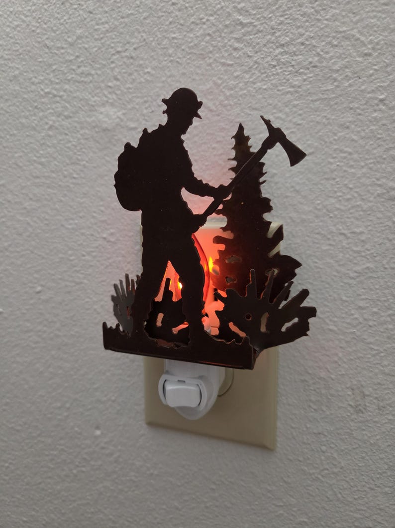 May include: A dark brown metal night light featuring a silhouette of a firefighter holding an axe, standing in front of a pine tree. The light emits a warm orange glow from behind the metal cutout, which is plugged into a standard wall outlet.