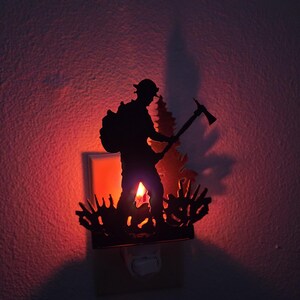 May include: A black metal night light featuring a silhouette of a firefighter carrying a backpack and holding an axe, standing in front of a pine tree and low brush. The light emits a warm red glow from behind the cutout metal design.