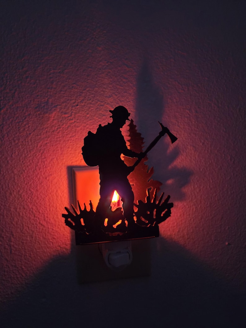 May include: A black metal night light featuring a silhouette of a firefighter carrying a backpack and holding an axe, standing in front of a pine tree and low brush. The light emits a warm red glow from behind the cutout metal design.
