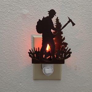 May include: A dark brown metal night light featuring a silhouette of a firefighter wearing a helmet and carrying an axe, standing in front of a pine tree. The light emits a warm orange glow from behind the metal cutout, illuminating the wall.