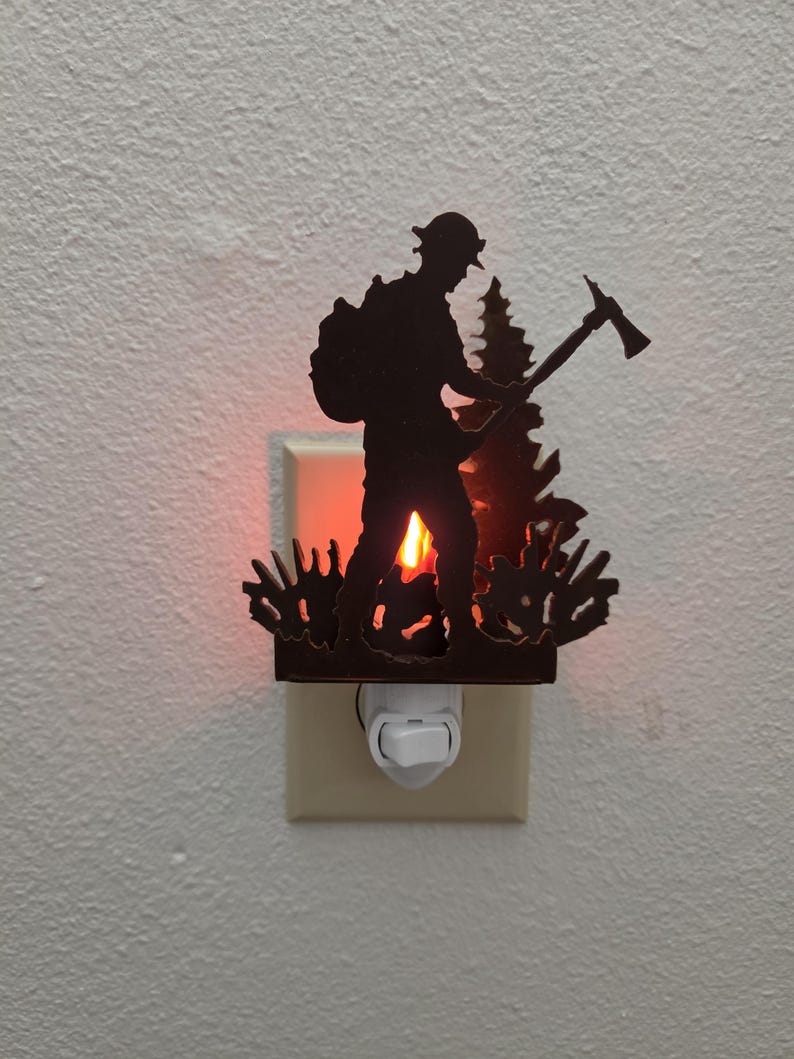 May include: A dark brown metal night light featuring a silhouette of a firefighter wearing a helmet and carrying an axe, standing in front of a pine tree. The light emits a warm orange glow from behind the metal cutout, illuminating the wall.