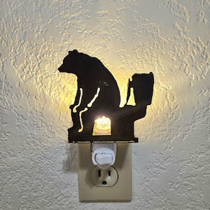 May include: Black metal night light featuring a bear silhouette seated on a toilet. The night light plugs into a standard wall outlet and emits a warm, yellow glow. The bear is depicted in a humorous pose, adding a touch of whimsy to the decor.
