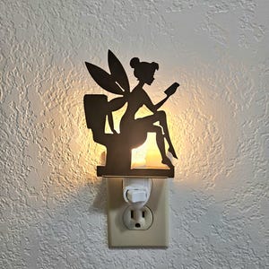 May include: A decorative night light featuring a silhouette of a fairy reading a book while seated. The fairy is black and has large wings. The night light plugs into a standard wall outlet and emits a soft, warm glow.