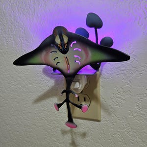 May include: A black metal night light shaped like a manta ray with pink and green accents. The light is plugged into an electrical outlet and emits a purple glow.