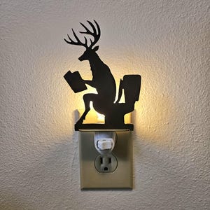 May include: A black silhouette night light featuring a deer with antlers sitting on a toilet while reading a book. The plug-in light fixture emits a warm glow from behind the cutout design, which is mounted directly into a standard wall electrical outlet.