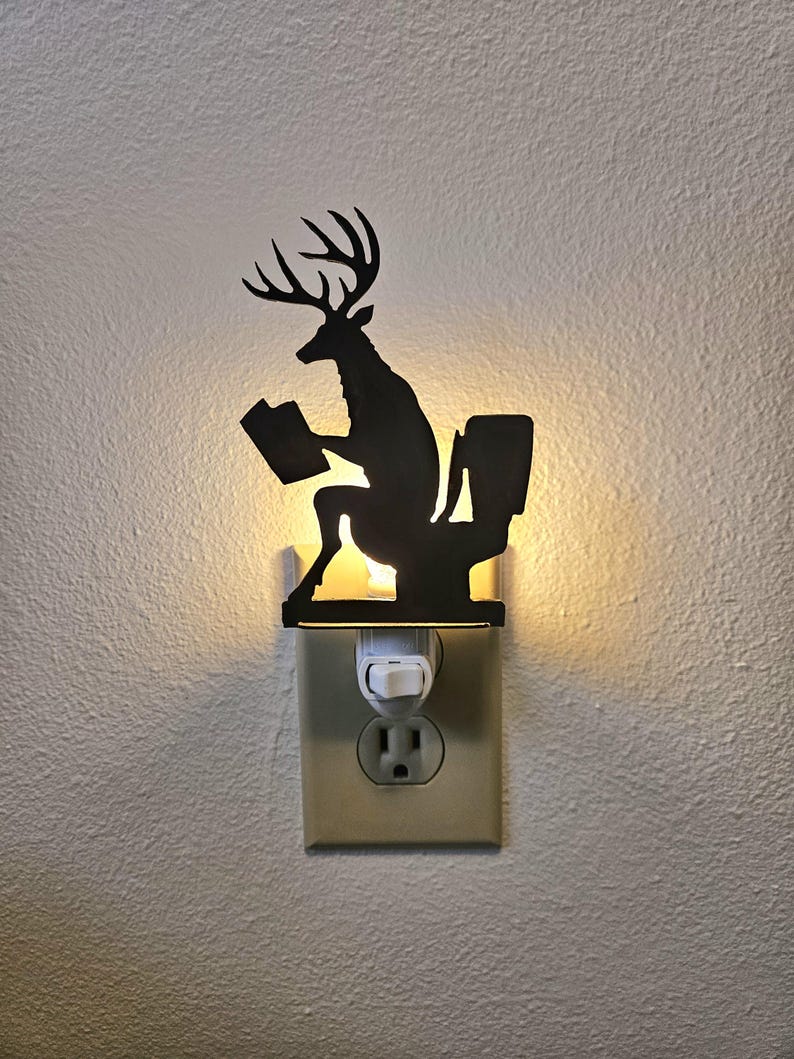 May include: A black silhouette night light featuring a deer with antlers sitting on a toilet while reading a book. The plug-in light fixture emits a warm glow from behind the cutout design, which is mounted directly into a standard wall electrical outlet.