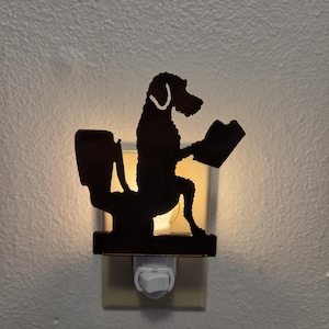 May include: A black nightlight featuring a silhouette of a dog sitting on a toilet while reading a book. The nightlight is plugged into a wall outlet and emits a warm, glowing light from behind the dark, cutout figure of the animal.