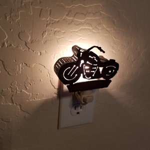 Könnte beinhalten: Ein dekoratives Nachtlicht in Form eines Motorrads. Die schwarze Metall-Silhouette des Motorrads ist in eine weiße Steckdose an einer strukturierten Wand eingesteckt. Das Licht scheint durch das Design des Motorrads.