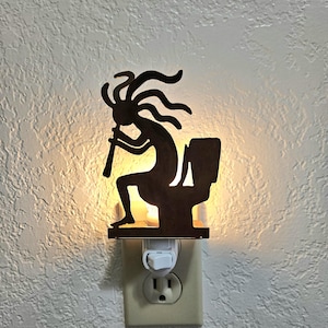 May include: A brown metal night light featuring a silhouette of Kokopelli playing a flute while seated on a toilet. The night light plugs into a standard wall outlet and emits a soft glow.