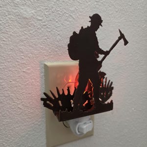May include: A dark brown night light featuring a silhouette of a firefighter carrying an axe, positioned in front of a glowing red light. The decorative piece is mounted onto a standard white electrical wall outlet cover with a toggle switch.