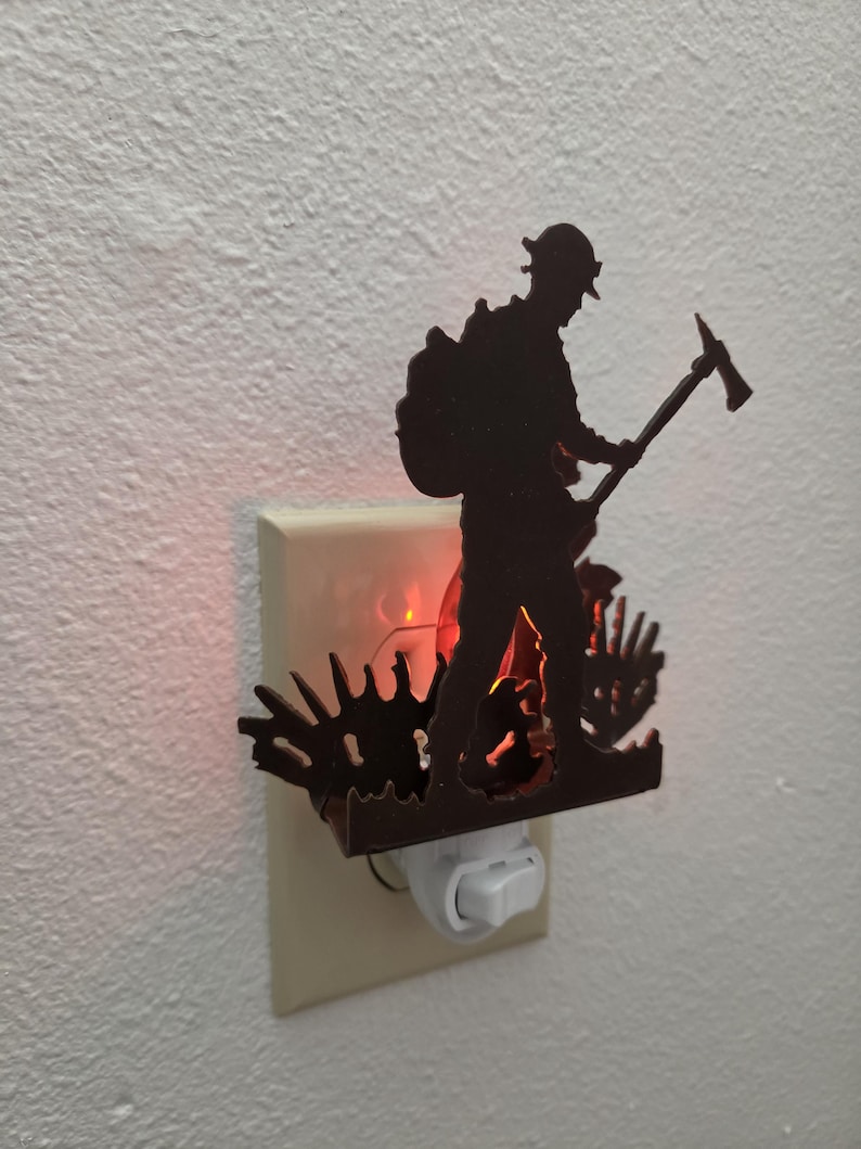 May include: A dark brown night light featuring a silhouette of a firefighter carrying an axe, positioned in front of a glowing red light. The decorative piece is mounted onto a standard white electrical wall outlet cover with a toggle switch.