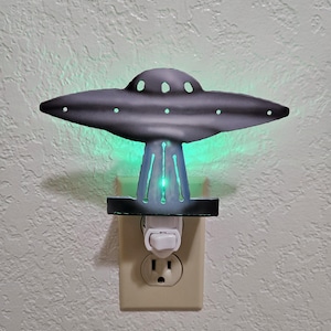 May include: A gray metal UFO night light with green internal lighting. The night light plugs directly into a standard wall outlet. The UFO has a classic saucer shape with multiple portholes.
