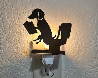 Pode incluir: Uma luz noturna de metal preto com a silhueta de um cão dachshund sentado numa sanita enquanto lê um livro. O dispositivo de ligar à tomada emite um brilho quente por trás da forma do cão, montada numa tomada de parede branca padrão.