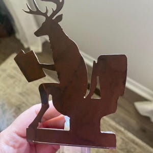 May include: A brown metal nightlight cover featuring a silhouette of a deer sitting on a toilet while reading a book. The deer has antlers and is positioned in profile. The metal cutout is mounted on a small plastic base with an off and on switch.