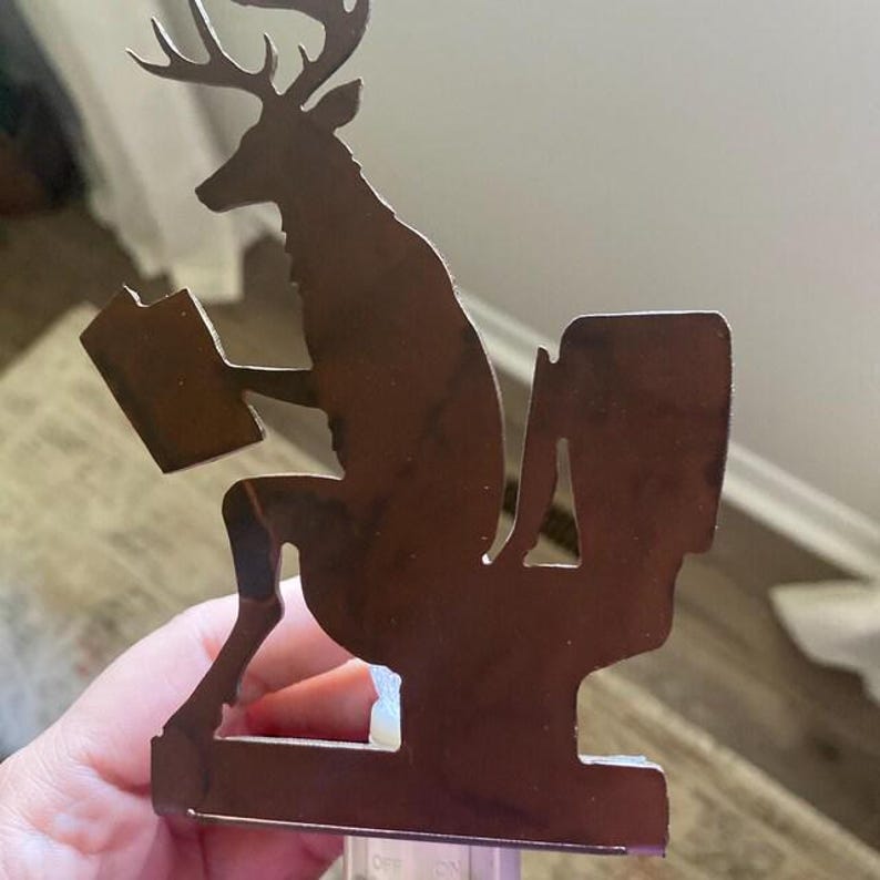May include: A brown metal nightlight cover featuring a silhouette of a deer sitting on a toilet while reading a book. The deer has antlers and is positioned in profile. The metal cutout is mounted on a small plastic base with an off and on switch.