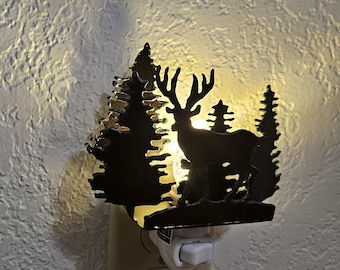 May include: A decorative night light featuring a silhouette of a deer and trees. The metal cutout design is black, with a warm light emanating from behind the deer. The night light plugs into a standard wall outlet.