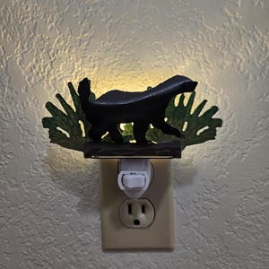 Può includere: Una luce notturna nera a forma di tasso, con dettagli di fogliame verde. Si collega direttamente a una presa elettrica standard ed emette una luce soffusa, ideale per camere da letto o corridoi.