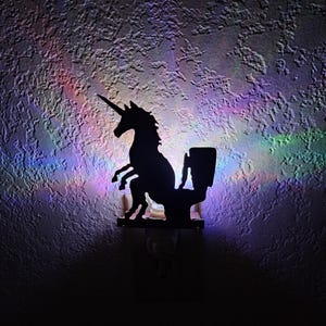 May include: A black silhouette night light depicting a unicorn on a toilet. The night light is fixed to a textured wall. The wall is lit with a spectrum of colourful light.