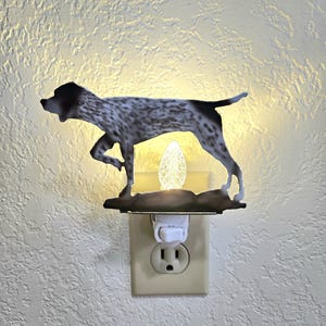Könnte beinhalten: Ein weißes Nachtlicht mit einer Metallsilhouette eines Deutsch Kurzhaar Hundes. Das Nachtlicht ist an eine normale Steckdose angeschlossen.