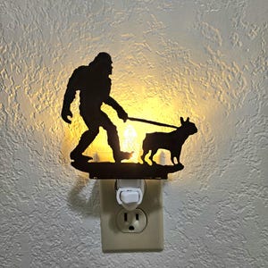 May include: A black metal night light featuring a silhouette of Bigfoot walking a French bulldog. The night light is plugged into a standard electrical outlet.