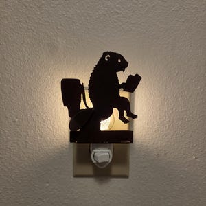 May include: A plug-in night light featuring a dark silhouette of a beaver sitting on a toilet while reading a book. The light emits a warm glow from behind the cutout figure, which is mounted on a standard white electrical wall outlet fixture.