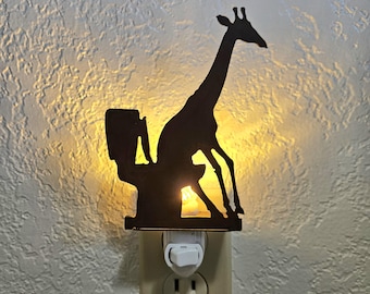 May include: A dark silhouette night light featuring a giraffe sitting on a toilet. The plug-in fixture emits a warm yellow glow from behind the cutout metal shape, casting light onto the wall surface while illuminating the giraffe's outline.